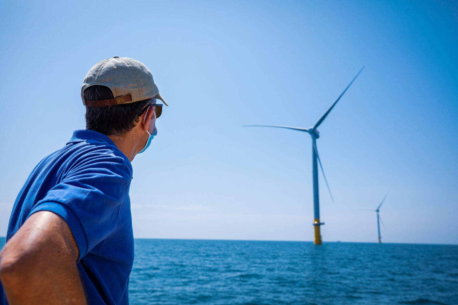 Man with mask and hat on looks out into body of water at wind farm